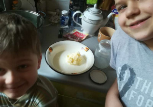 Dwóch uśmiechniętych chłopców a miedzy nimi miseczka z masłem wykonanym domowym sposobem.